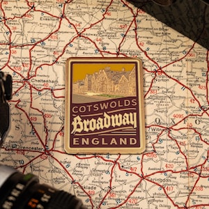 Może przedstawiać: Prostokątny magnes z Cotswolds Broadway w Anglii. Posiada stylizowany, zabytkowy design z ilustracją budynku i tekstem w kolorze bordowym i złotym na tle mapy.
