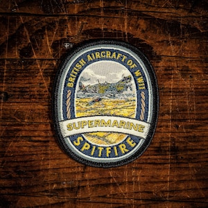 May include: An oval embroidered patch featuring a Supermarine Spitfire aircraft in flight over a landscape. The border is dark blue with gold rope detailing. Text reads BRITISH AIRCRAFT OF WWII, SUPERMARINE, and SPITFIRE in gold and white lettering.