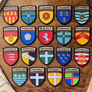 K&ouml;nnte beinhalten: Eine Sammlung von 20 bunten gestickten Aufn&auml;hern mit den Wappen verschiedener englischer Grafschaften. Jeder Aufn&auml;her hat die Form eines Schildes und zeigt ein einzigartiges Design, darunter Tiere, Pflanzen und geometrische Muster. Die Aufn&auml;her sind in einem Raster auf einer Holzoberfl&auml;che angeordnet.