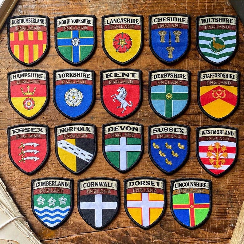 K&ouml;nnte beinhalten: Eine Sammlung von 20 bunten gestickten Aufn&auml;hern mit den Wappen verschiedener englischer Grafschaften. Jeder Aufn&auml;her hat die Form eines Schildes und zeigt ein einzigartiges Design, darunter Tiere, Pflanzen und geometrische Muster. Die Aufn&auml;her sind in einem Raster auf einer Holzoberfl&auml;che angeordnet.