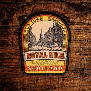 May include: Embroidered patch depicting a street scene, with the words "OLD TOWN EDINBURGH" and "ROYAL MILE SCOTLAND". The patch has a black border and is set against a textured wooden background. The design is in shades of orange, yellow, and black.
