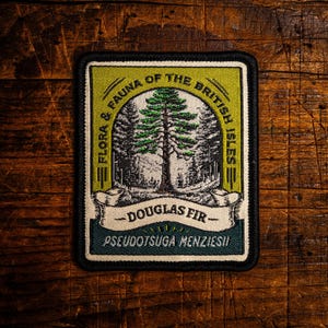 Op de afbeelding: Een rechthoekige patch met een zwarte rand toont een gedetailleerde illustratie van een Douglas spar in een boslandschap. De tekst "FLORA & FAUNA OF THE BRITISH ISLES" staat in een boog bovenaan, met "DOUGLAS FIR" en "PSEUDOTSUGA MENZIESII" eronder.