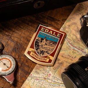 May include: A rectangular metal sign with the text "EDALE PEAK DISTRICT DERBYSHIRE" in red, gold, and blue. The sign is on a wooden surface with a map, compass, and camera.