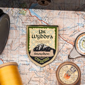 Puede incluir: Un pin de esmalte dorado y verde con un diseño de montaña y el texto "Yr Wyddfa 1085m Snowdon Cymru".