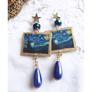 Puede incluir: Pendientes colgantes con una reproducción en miniatura de "La noche estrellada" de Van Gogh. Tienen marcos dorados, detalles de estrellas y cuentas azules facetadas, y colgantes azul marino en forma de lágrima. Se exhiben sobre una superficie blanca texturizada.