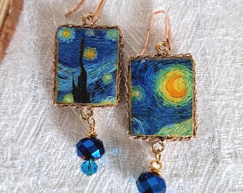 May include: A pair of earrings featuring rectangular pendants with a painting of Van Gogh's "Starry Night." The pendants are framed in gold-colored metal and dangle with blue crystal beads. The earrings have gold-colored ear wires.