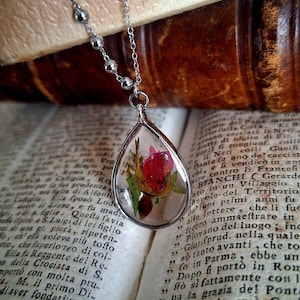 May include: Silver teardrop pendant necklace with a real red rose bud encased in clear resin. The pendant hangs from a delicate silver chain with small beaded accents. The necklace rests on the pages of an open antique book.