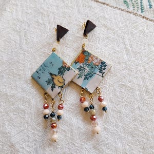 May include: A pair of dangle earrings featuring a Japanese-inspired design. Each earring has a dark brown triangle stud, a diamond-shaped pendant with a bird and floral print, and beaded dangles in shades of red, white, and black.