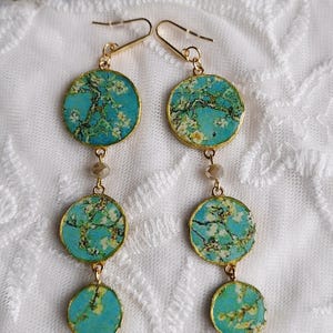 Könnte beinhalten: Ohrringe zum Abhängen mit einem türkis-goldenen Design. Jeder Ohrring hat drei kreisförmige Anhänger mit einem Blumenmuster, die durch kleine Perlen verbunden sind. Die Ohrringe haben goldfarbene Haken.