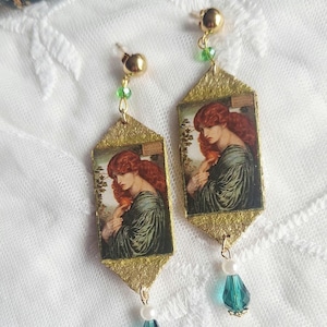 May include: A pair of drop earrings featuring rectangular pendants with a printed portrait of a red-haired person in a green robe. The pendants have a textured gold-tone border, accented with small green beads, white pearls, and teal teardrop crystals.