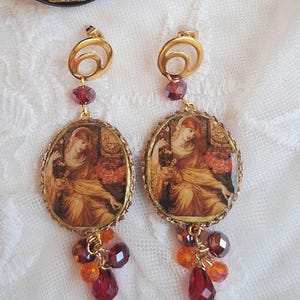 May include: Gold-toned dangle earrings featuring oval pendants with a Renaissance-style painting of a woman in a yellow gown. The earrings have red and orange bead clusters and a pearl drop. The top has a gold swirl design.