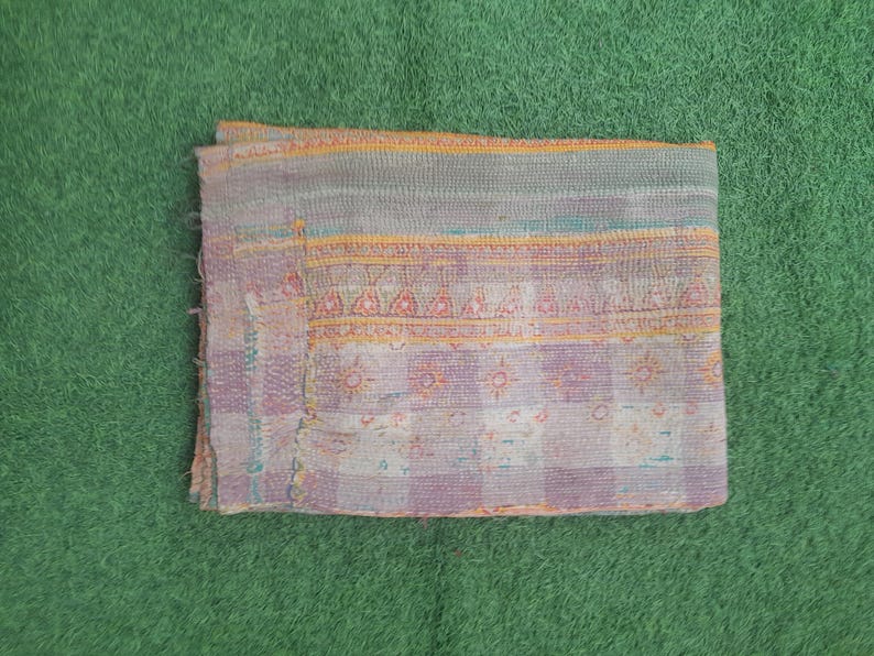 Puede incluir: Textil rectangular plegado con un dise&ntilde;o de patchwork. La tela presenta una mezcla de colores, incluyendo lavanda, azul p&aacute;lido y amarillo, con un patr&oacute;n de cuadros. El textil est&aacute; colocado sobre una superficie verde.