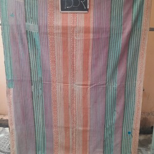 Puede incluir: Textil a rayas colorido con bandas verticales de color naranja, morado y turquesa. El textil tiene un aspecto vintage descolorido, con costuras visibles y un borde decorativo. Una peque&ntilde;a pizarra con el n&uacute;mero "352" est&aacute; sujeta en la parte superior.