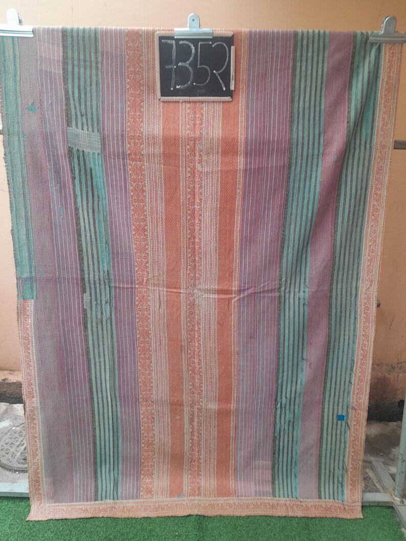 Puede incluir: Textil a rayas colorido con bandas verticales de color naranja, morado y turquesa. El textil tiene un aspecto vintage descolorido, con costuras visibles y un borde decorativo. Una peque&ntilde;a pizarra con el n&uacute;mero "352" est&aacute; sujeta en la parte superior.