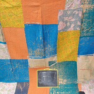 May include: A colorful patchwork quilt with a variety of fabrics and patterns. The quilt is made up of squares and rectangles of different colors, including blue, orange, yellow, and white. The quilt is folded in half and has a small chalkboard with the number 6757 written on it.