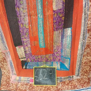 May include: A colorful patchwork quilt with a variety of fabrics and textures. The quilt is made up of squares and rectangles of different colors and patterns. The quilt is edged with a black border. A small black chalkboard with the number 6629 written on it is placed on the quilt.