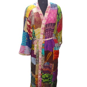Op de afbeelding: Een lange, kleurrijke patchwork-jas met een strikceintuur. De jas is gemaakt van verschillende stoffen in tinten roze, paars, groen, blauw en bruin.