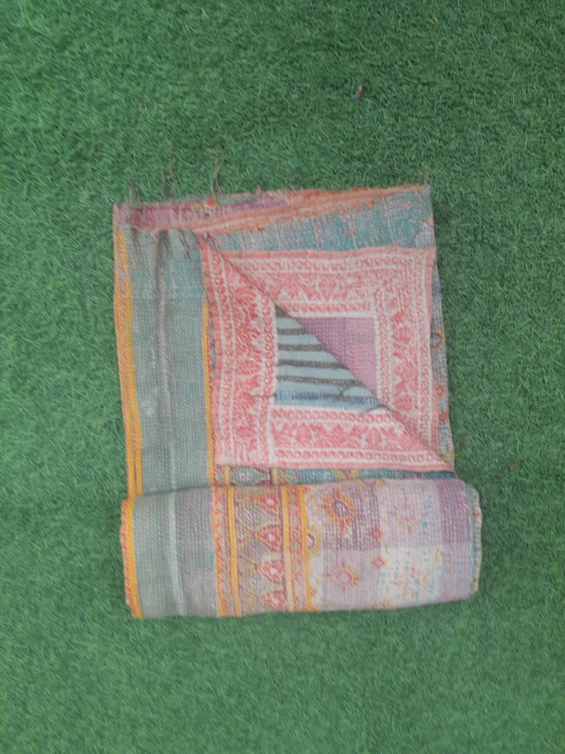 Puede incluir: Colcha patchwork plegada con una mezcla de colores que incluyen turquesa, rosa y morado. La colcha presenta intrincados patrones y dise&ntilde;os, con una secci&oacute;n enrollada en la parte inferior. La colcha est&aacute; colocada sobre una superficie verde.