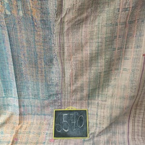May include: A large, textured textile with a patchwork design. The fabric features a blend of pastel colors, including blue, pink, and beige, with visible stitching. A small chalkboard with the number "6570" is in the foreground.