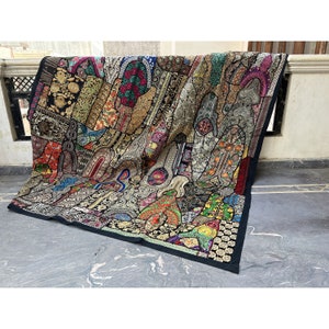 Op de afbeelding: Een groot, kleurrijk patchworktextiel met een zwarte rand. Het textiel heeft een verscheidenheid aan patronen en kleuren, waaronder goud, rood, groen en blauw. Het textiel is over een richel gedrapeerd.