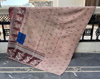 Op de afbeelding: Een roze en bruine kantha-deken met een bloemenpatroon en een blauwe patch. De deken is gevouwen en over een leuning gelegd.