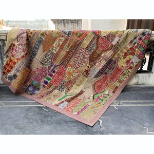 Può includere: Una colorata trapunta patchwork con una varietà di tessuti fantasia nei toni del rosso, oro, verde e viola. La trapunta ha un bordo rosa ed è drappeggiata su una sporgenza di pietra. Il design è un mix di motivi floreali e geometrici.