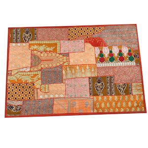 Op de afbeelding: Een rechthoekige patchwork wandhanger met een rode rand. De patchwork is gemaakt van verschillende stoffen in tinten oranje, bruin, roze en wit. De stoffen zijn versierd met ingewikkelde borduursels en pailletten.