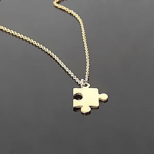 May include: A gold-colored necklace with a puzzle piece pendant. The chain is delicate, and the puzzle piece is a simple, elegant design. The necklace is displayed on a dark, reflective surface, with a few decorative, light-colored stones.