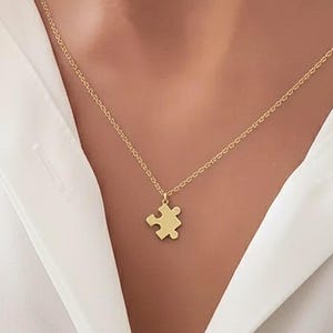 Può includere: Una collana color oro con un piccolo ciondolo a forma di pezzo di puzzle. Il ciondolo pende da una catena delicata. La collana è indossata su una camicia bianca.