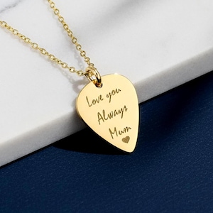 May include: A gold-colored pendant necklace with a heart-shaped charm. The charm is engraved with the words "Love you Always Mum" and a small heart symbol. The necklace is displayed on a white marble surface against a dark blue background.