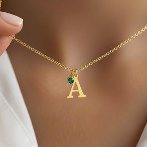 以下が含まれることがあります： 細いチェーンと、大文字の「A」のチャーム、そして小さな丸い深緑色の宝石のチャームが組み合わさったペンダントトップが付いたゴールドトーンのネックレス。金属部分は光沢のある明るいゴールドカラーで仕上げられています。