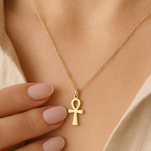 May include: A gold-colored Ankh pendant necklace. The pendant is a stylized cross with a loop at the top, suspended from a delicate chain. The necklace is worn against a neutral-toned shirt.