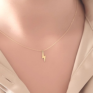 Könnte beinhalten: Eine zarte goldene Halskette mit einem kleinen Blitz-Anhänger. Der Anhänger hängt an einer feinen Kette. Die Halskette wird auf einem neutralfarbenen Hemd getragen. Ein schlichtes, stilvolles Accessoire.