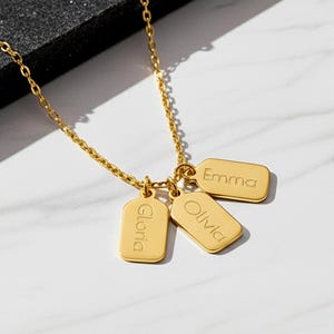 Op de afbeelding: Een goudkleurige ketting met een fijne schakelketting en drie kleine, rechthoekige hangertjes. Op elk plaatje is een naam gegraveerd: Emma, Olivia en Gloria. Het metalen oppervlak heeft een gepolijste afwerking en de hangertjes hangen samen.