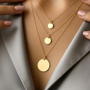 Può includere: Un set di tre collane d'oro a strati, ciascuna con una catena sottile e un ciondolo circolare. I ciondoli sono di diverse dimensioni, creando un effetto a strati. I gioielli sono mostrati su uno sfondo neutro, evidenziando il colore caldo dell'oro.