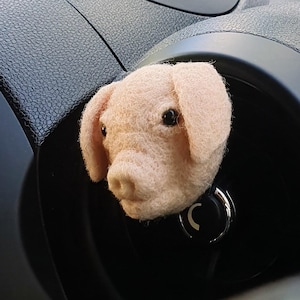 May include: A pink felt pig's head car charm with black eyes and a small snout. The charm is attached to a black car dashboard.