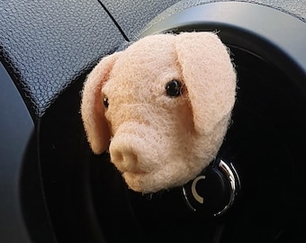 May include: A pink felt pig head car charm with black eyes and a small snout. The charm is attached to a black car dashboard.