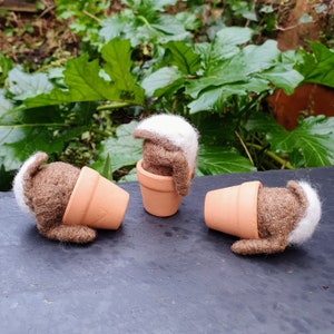 May include: Three handcrafted felt figures, each with a brown body and a white head, are arranged with terracotta plant pots. One figure stands in a pot, while the others are partially inside or tipped over. The background shows green foliage.