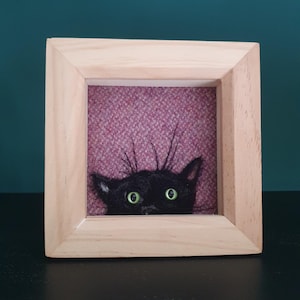 May include: A square light-colored wooden frame containing a needle-felted black cat head with bright green eyes. The cat is positioned against a textured, dark pink fabric background, with its ears and whiskers visible at the bottom of the frame.