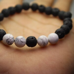 May include: A stretch bracelet composed of round, porous black lava beads interspersed with smooth, white marble-like beads featuring grey veining. The design alternates between the textured black stones and the polished white stones in a circular pattern.