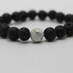 May include: A stretch bracelet composed of numerous round, porous black lava beads arranged in a circular pattern, featuring a single smooth, round white bead with grey veining positioned as a central accent piece within the bead sequence.