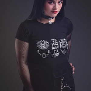 Puede incluir: Camiseta negra con el texto "IT'S VERY RUDE TO STARE" y dos diseños gráficos blancos. La modelo lleva una gargantilla negra con un candado plateado en forma de corazón. La modelo también lleva mangas de red negras.
