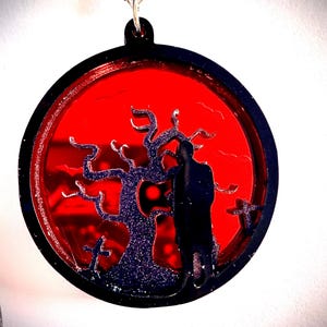 May include: A circular black pendant with a red backdrop depicting a spooky scene. A silhouetted figure stands near a tree and crosses. The pendant is suspended from a silver chain.