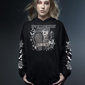 Op de afbeelding: Zwarte hoodie met een afbeelding van een skelet in een kist, vleermuizen en spinnenwebben. De tekst luidt "IT'S TOO PEOPLEY OUTSIDE" en "BACK TO BED". De mouwen hebben witte spinnenwebben en vleermuizen.