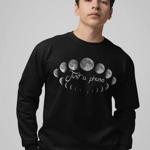 May include: Black sweatshirt featuring a graphic of the moon phases in shades of gray. The text "Just a phase" is written in a cursive font. The sweatshirt is a crew neck style.
