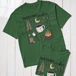 May include: Two green t-shirts with a nature-themed graphic. The design includes a pine tree, crescent moon, coffee mug, campfire, and the text "NATUREseeker". The shirts are displayed on a white wooden surface.