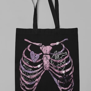 Puede incluir: Bolso tote negro con un dise&ntilde;o de caja tor&aacute;cica rosa y morado. La caja tor&aacute;cica tiene un coraz&oacute;n con un dise&ntilde;o cosido y una ara&ntilde;a. El bolso tiene dos asas negras.