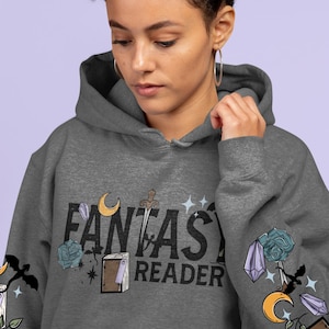 May include: A grey hoodie featuring the words "FANTASY READER" in black. The design incorporates a sword, crescent moon, book, and floral and crystal illustrations. The sleeves have additional fantasy-themed graphics.