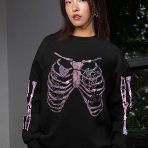 Può includere: Felpa nera con un design a gabbia toracica rosa e viola. La grafica include un cuore con la scritta "HEART" e un ragno. Le maniche hanno un design a ossa abbinato. Un top casual.