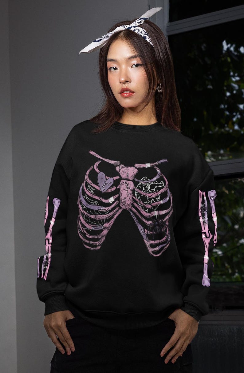 Puede incluir: Sudadera negra con dise&ntilde;o de caja tor&aacute;cica rosa y morada. El gr&aacute;fico incluye un coraz&oacute;n con la palabra "HEART" y una ara&ntilde;a. Las mangas tienen dise&ntilde;os de huesos a juego. Se lleva un pa&ntilde;uelo blanco y negro en la cabeza.